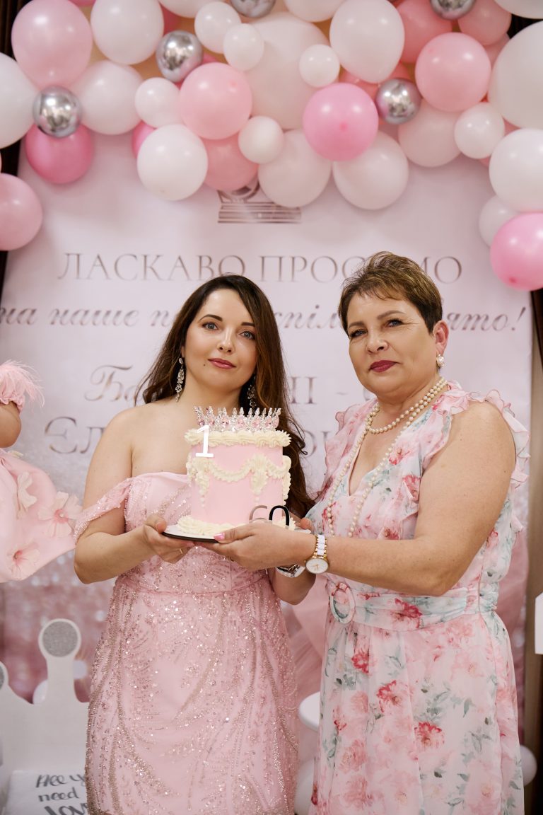 Дві жінки у рожевому одязі разом тримають святковий торт перед фотозоною
