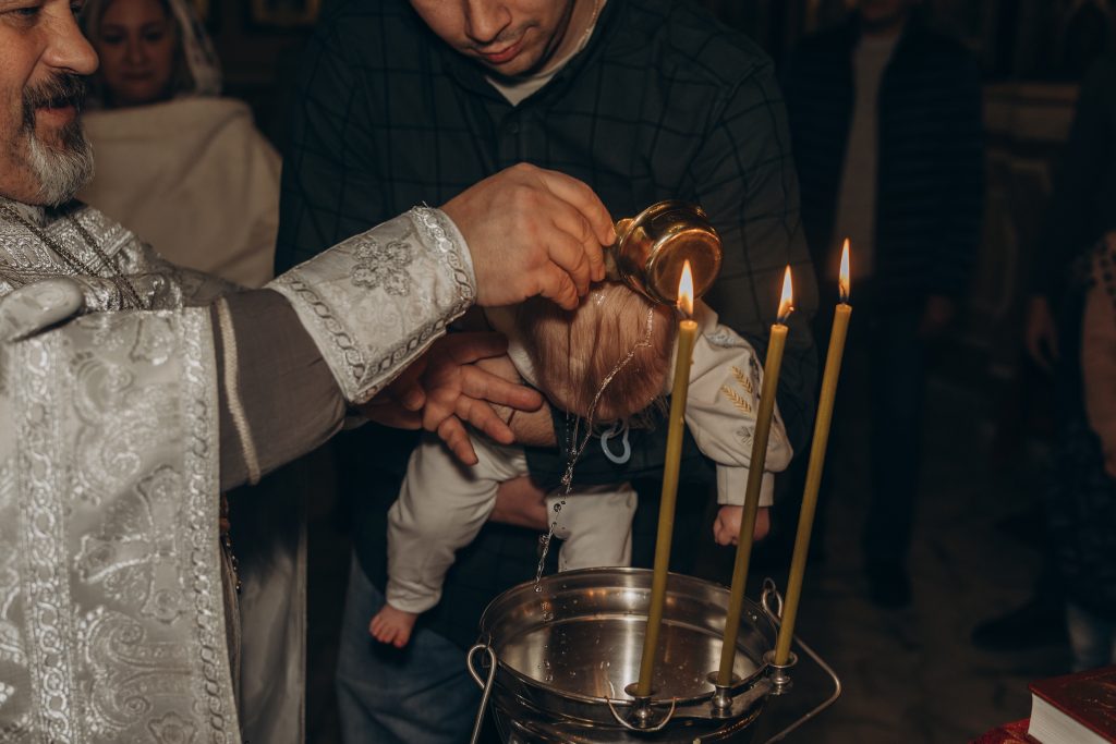 Священник поливає голову Олександра водою над купіллю, поки чоловік тримає дитину