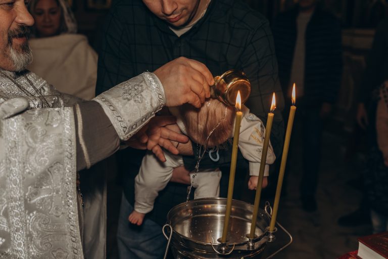 Священник поливає голову Олександра водою над купіллю, поки чоловік тримає дитину