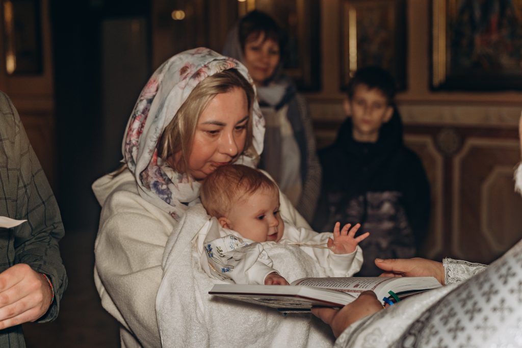 Жінка у світлій хустці тримає Олександра перед відкритою богослужбовою книгою