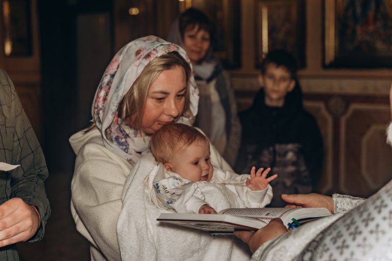 Жінка у світлій хустці тримає Олександра перед відкритою богослужбовою книгою