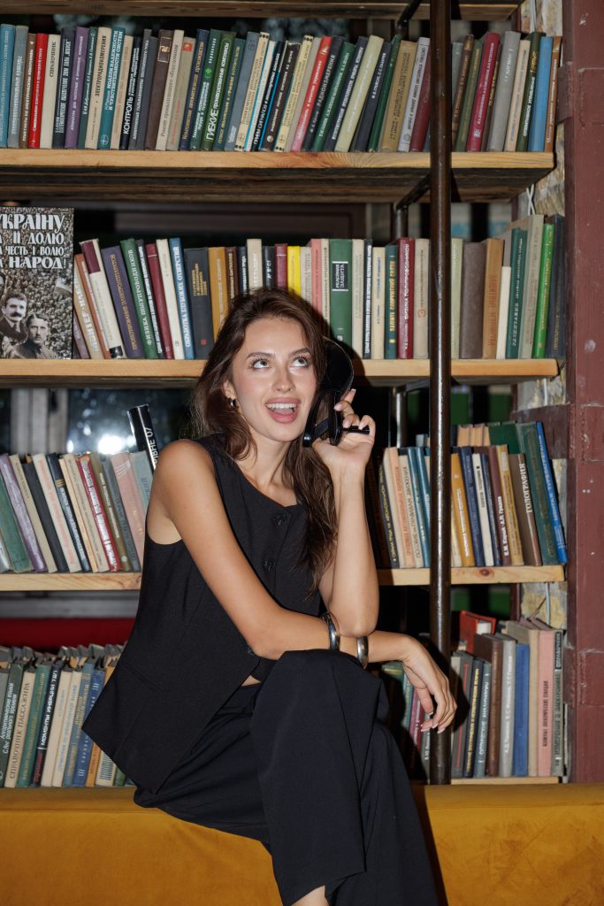 Усміхнена Даша тримає чорний туфель біля вуха на тлі книжкових полиць
