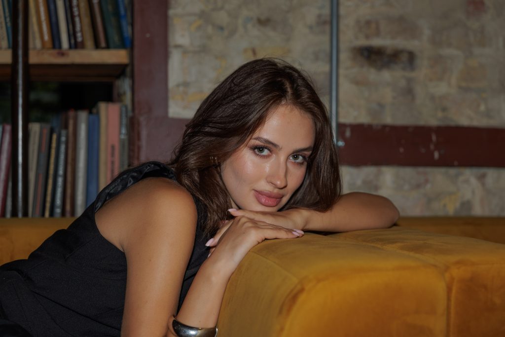 Портрет Даші у чорному жилеті біля гірчичного дивана та книжкових полиць