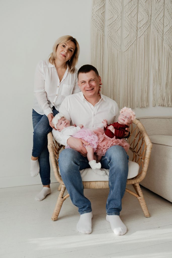 Сімейний портрет батьків із двома немовлятами у світлій студії біля плетеного крісла