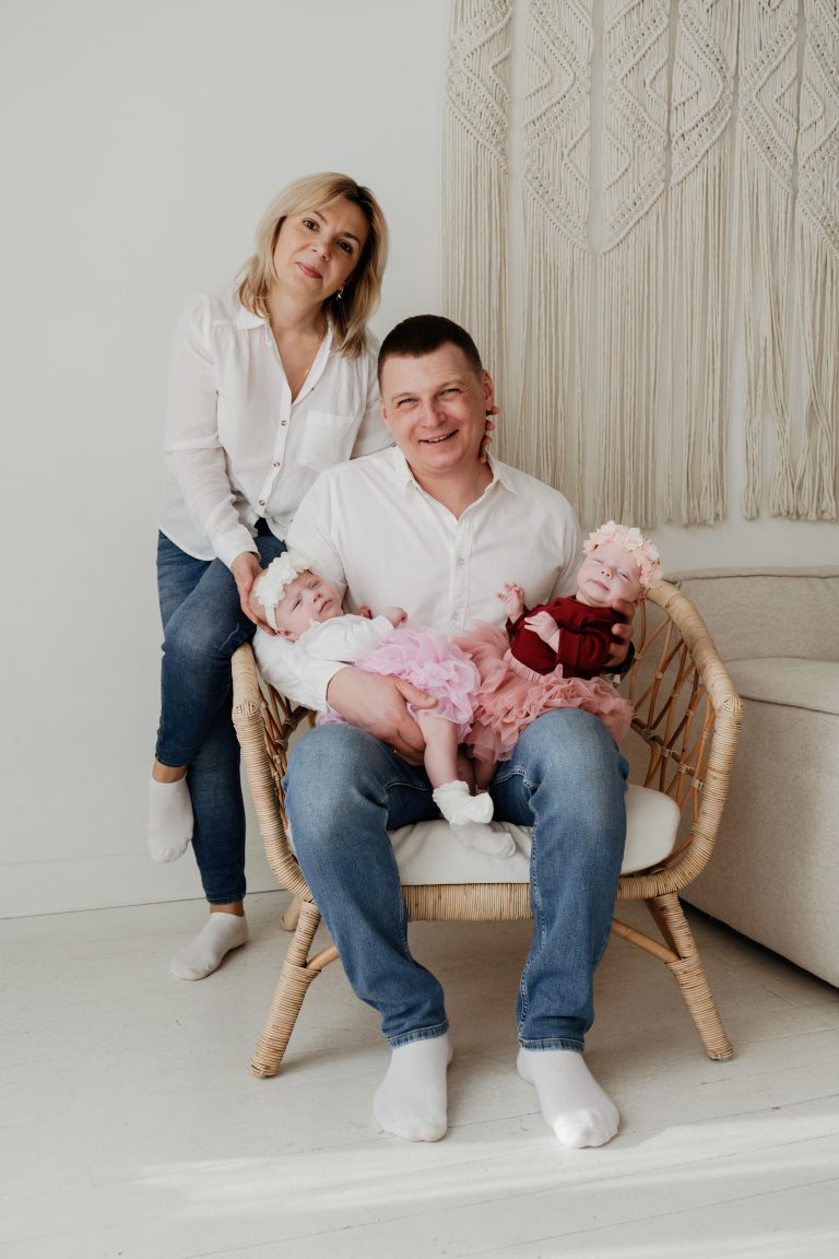 Сімейний портрет батьків із двома немовлятами у світлій студії біля плетеного крісла
