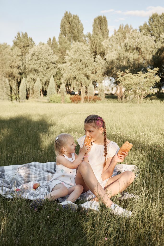 Дві дівчинки сидять на пледі у траві та діляться випічкою під час пікніка