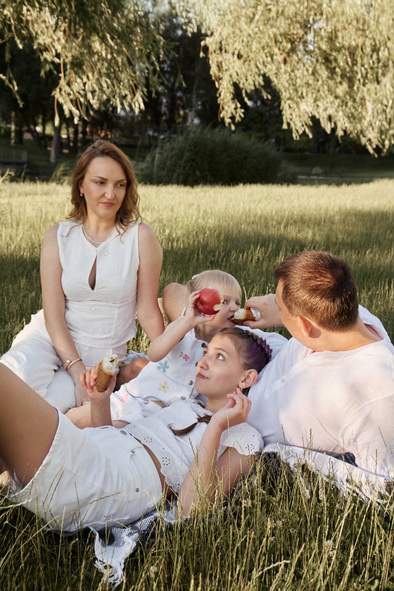 Родина відпочиває на пледі у високій траві та їсть яблуко і випічку