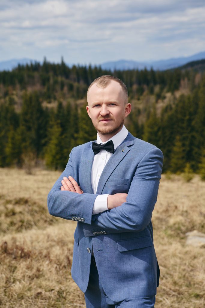 Портрет нареченого у блакитному костюмі з чорним метеликом на тлі гірського лісу.