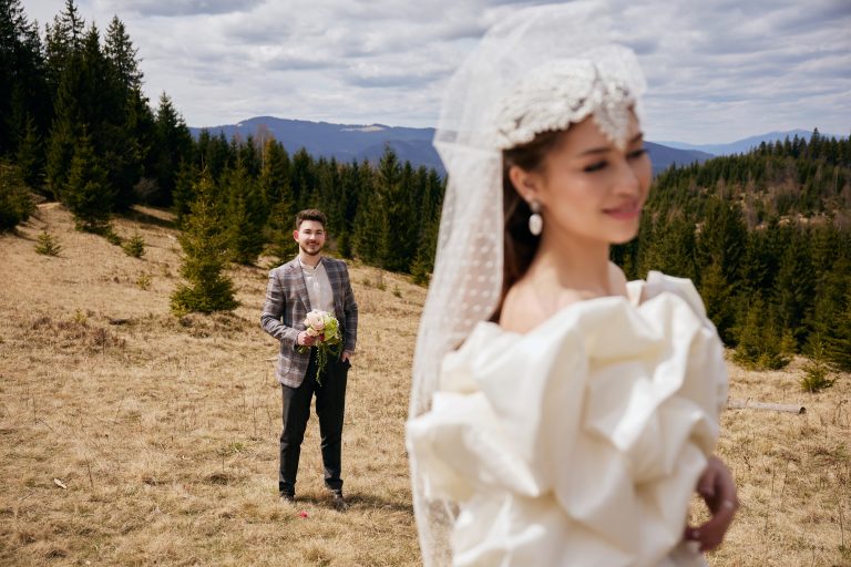 Наречений з букетом стоїть у фокусі на гірській галявині, а наречена у білій сукні та фаті розмита на передньому плані.