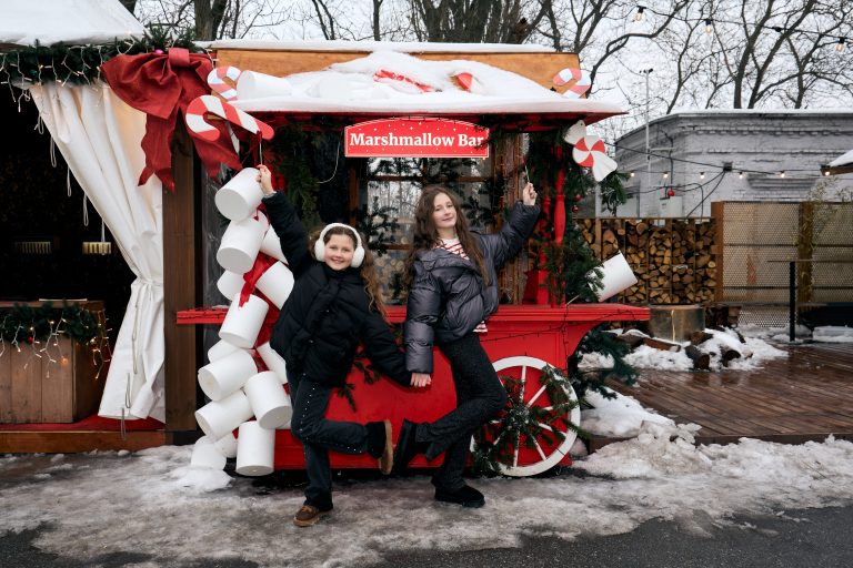 Дві дівчини позують біля червоного святкового кіоску Marshmallow Bar зі снігом і декораціями