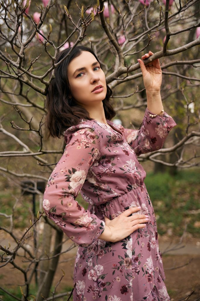 Ірина у рожевій квітковій сукні позує серед гілок магнолії без розкритих квітів