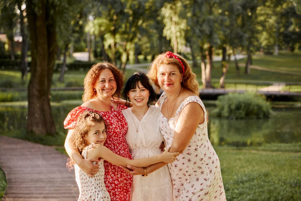 Чотири учасниці сімейної фотосесії стоять разом і обіймаються на тлі зеленої водойми в парку.