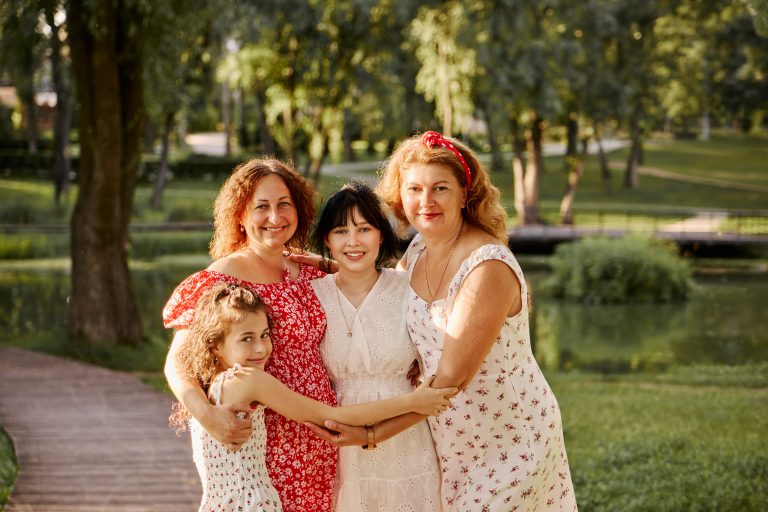 Чотири учасниці сімейної фотосесії стоять разом і обіймаються на тлі зеленої водойми в парку.