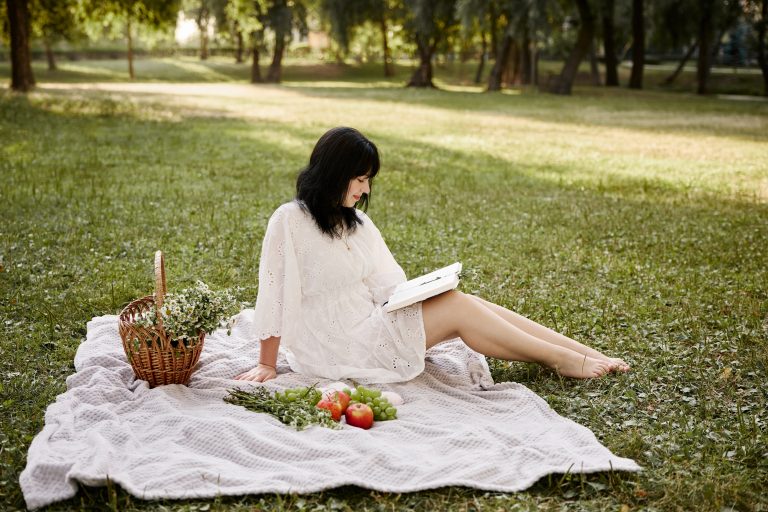 Аліса сидить на світлому пледі з відкритою книжкою, фруктами та квітами в парку