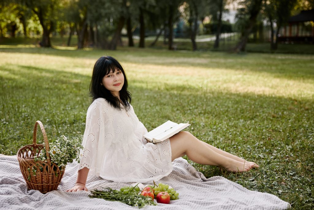 Аліса сидить на пледі з відкритою книжкою поруч із кошиком ромашок та фруктами