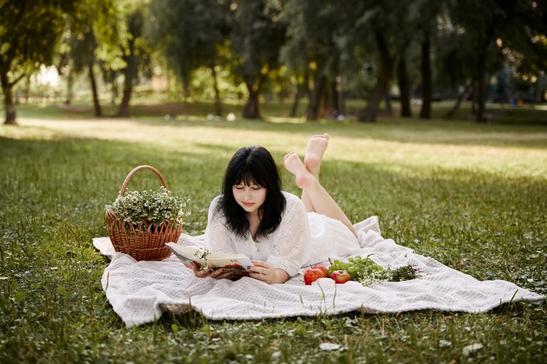 Аліса лежить на пледі й читає книжку поруч із кошиком квітів, яблуками та виноградом