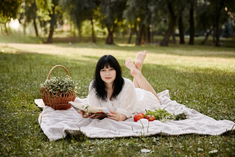 Аліса лежить на пледі з книжкою та дивиться в камеру, поруч кошик польових квітів і фрукти