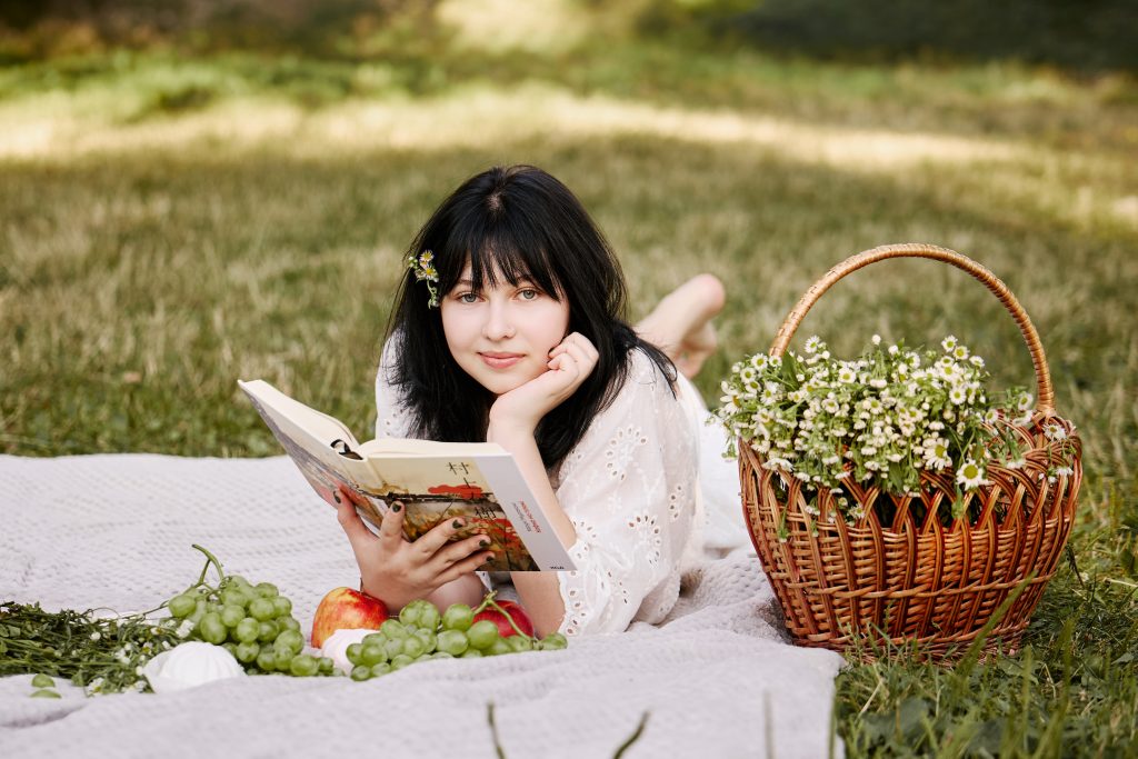 Аліса лежить на пледі з відкритою книжкою поруч із кошиком ромашок, яблуками та виноградом