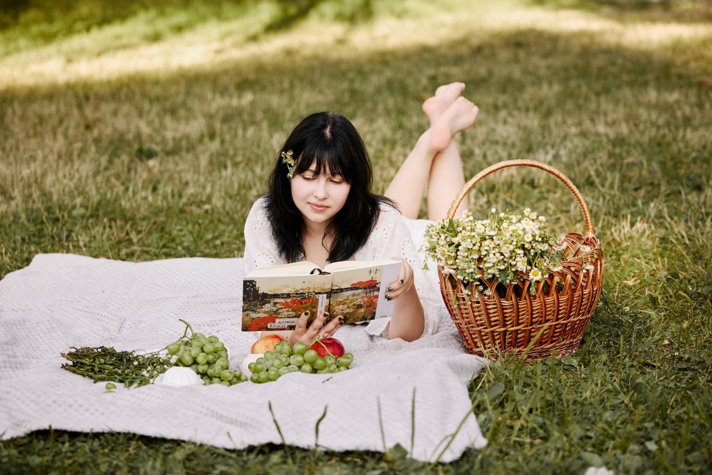 Аліса читає книжку лежачи на пледі поруч із великим кошиком білих польових квітів