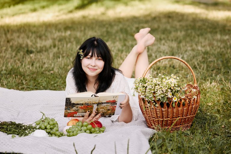 Аліса усміхається з відкритою книжкою, лежачи на пледі поруч із кошиком ромашок і фруктами