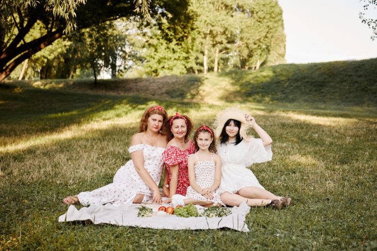 Чотири учасниці сімейної фотосесії сидять разом на пледі в зеленому парку серед фруктів і польових квітів.