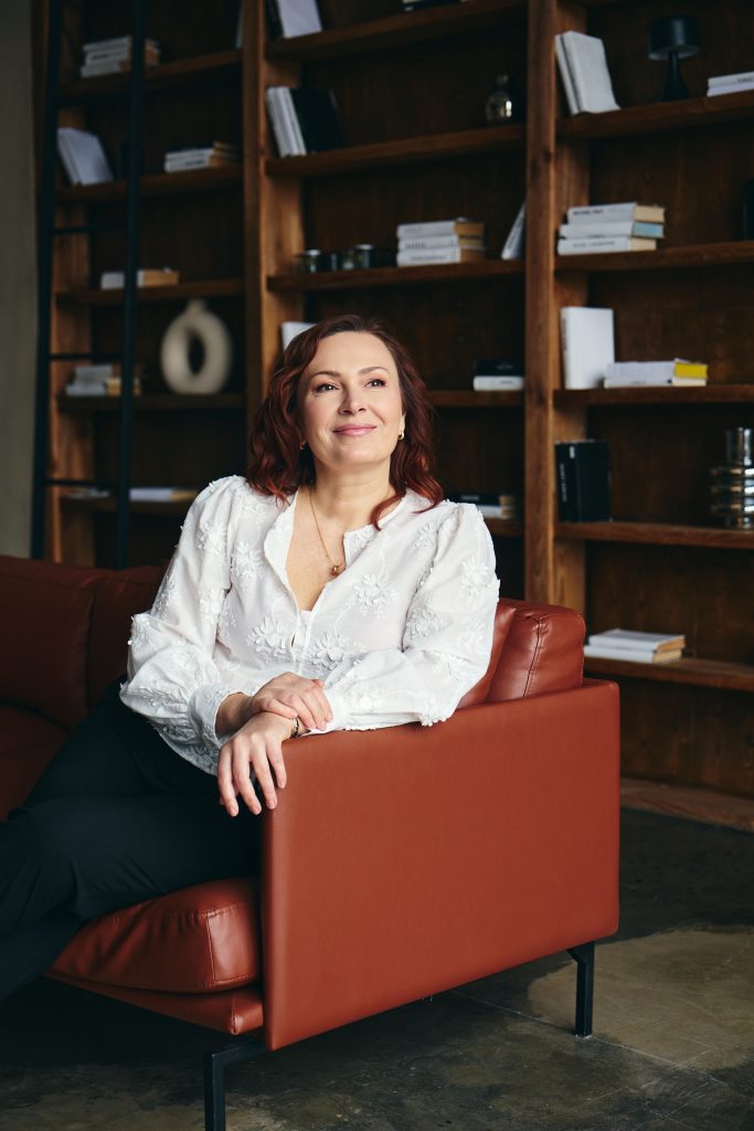Жінка в білій блузі сидить у кріслі на тлі дерев'яних книжкових полиць