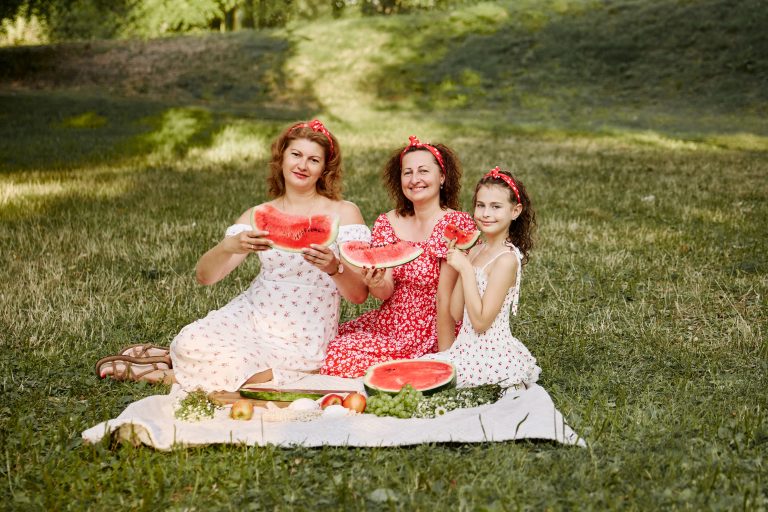 Три учасниці фотосесії сидять на пледі в парку та тримають великі скибки кавуна перед камерою.