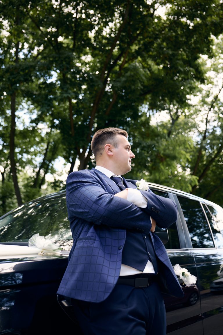 Максим позує у синьому костюмі біля темного весільного автомобіля