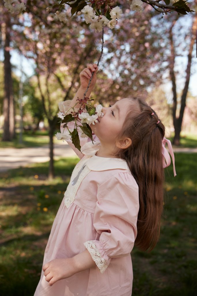Дівчинка у рожевій сукні нахилилася до гілки з білими весняними квітами