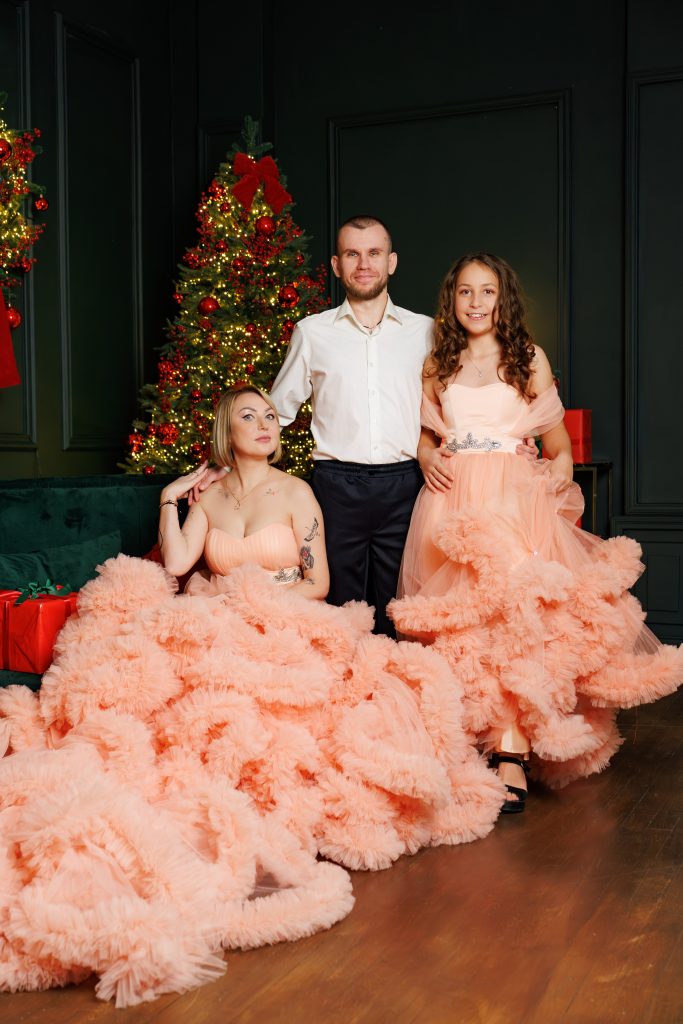 Валентина, жінка у персиковій сукні та чоловік у білій сорочці позують разом біля прикрашеної новорічної ялинки