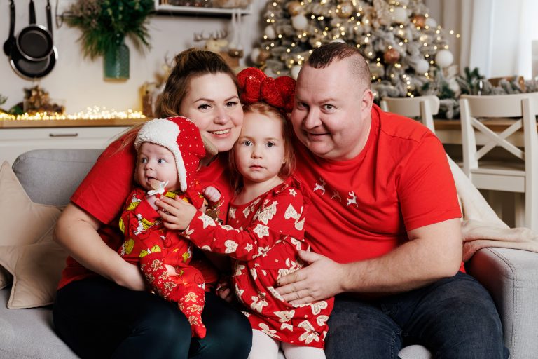 Дівчинка у червоній сукні та блискучому обручі з бантом лежить біля великих червоних подарункових коробок.