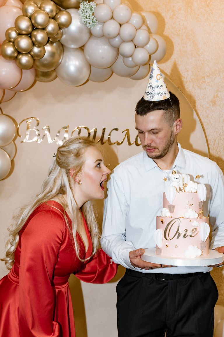 Жінка в червоній сукні та чоловік у святковому ковпаку тримають рожевий торт на фоні фотозони