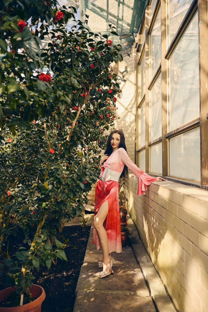 Жінка у рожево-червоній довгій сукні стоїть біля вікон серед квітучих кущів
