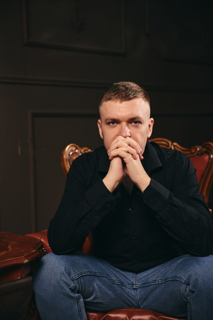 Кольоровий чоловічий портрет Дмитра в чорній сорочці, який сидить у кріслі зі складеними руками