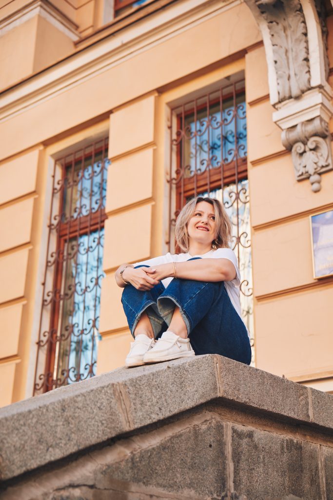 Марина сидить на кам'яному парапеті біля світлої історичної будівлі з декоративними вікнами.