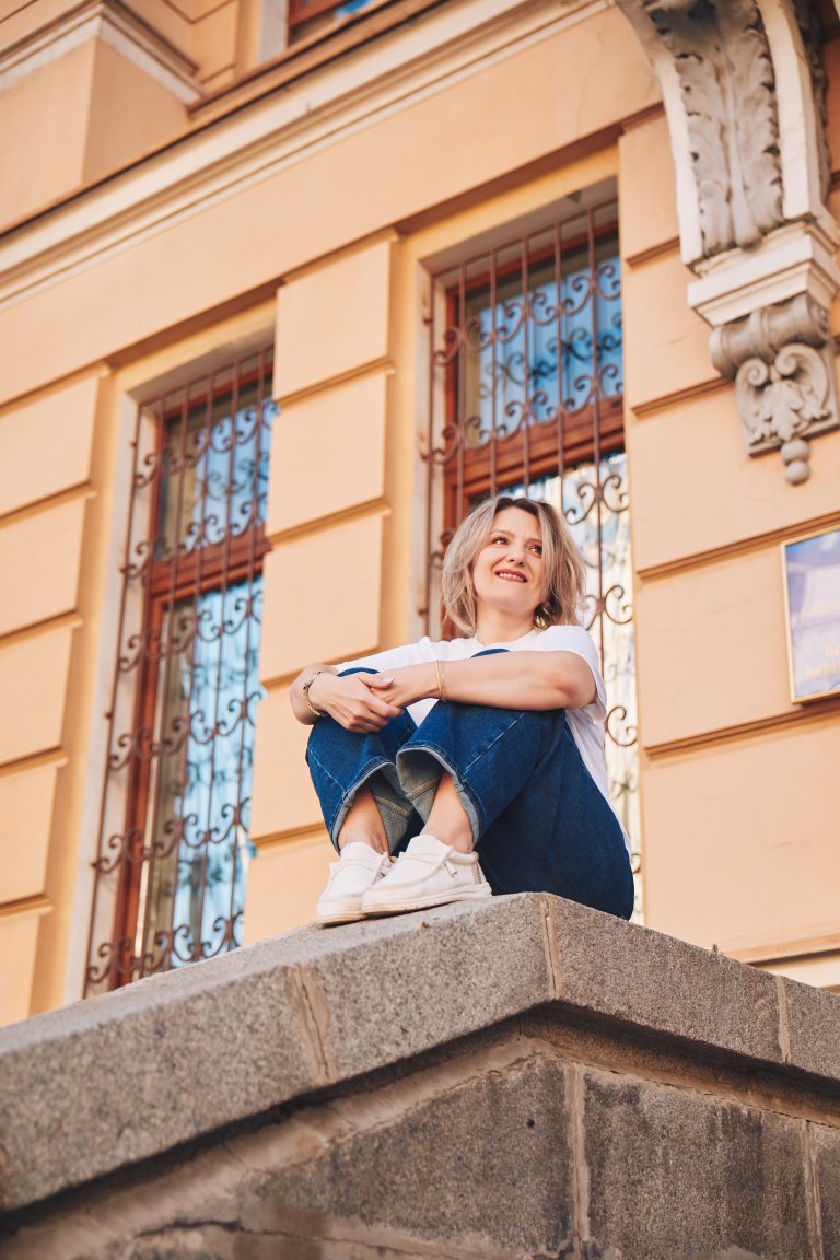 Марина сидить на кам'яному парапеті біля світлої історичної будівлі з декоративними вікнами.