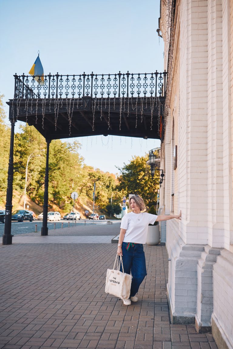 Марина стоїть біля світлої цегляної будівлі з великою текстильною сумкою, а над входом видно прапор України.
