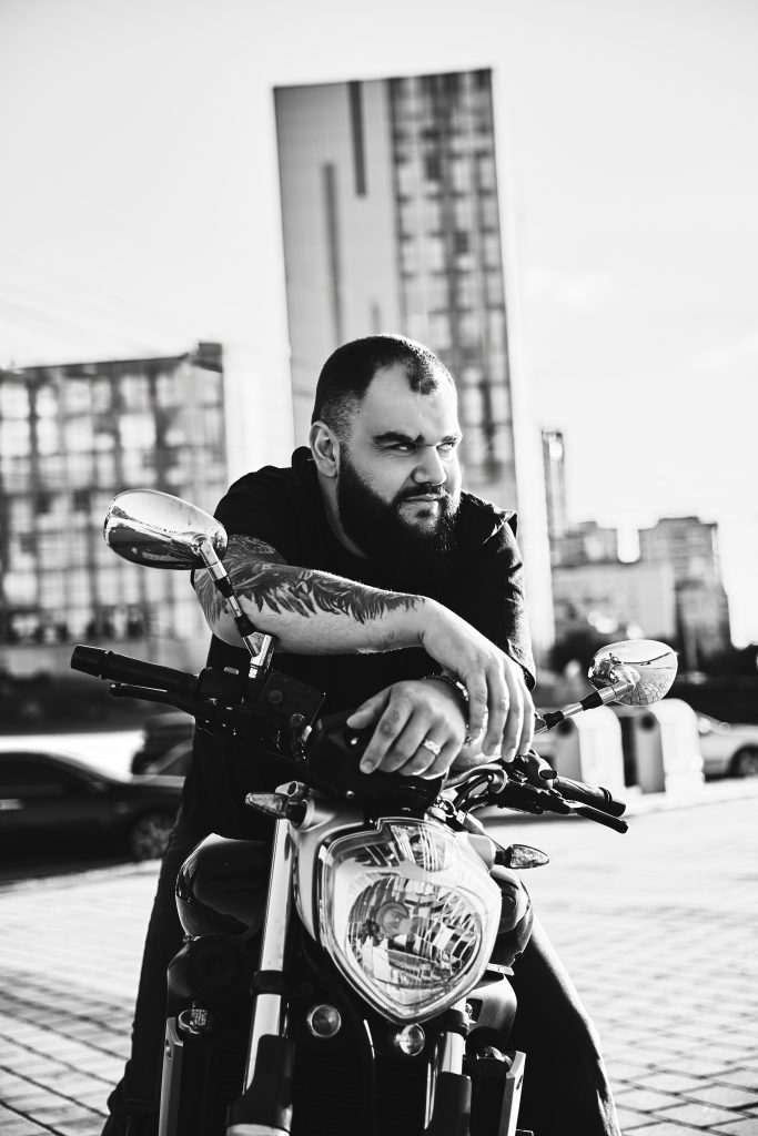 Чорно-білий портрет бородатого чоловіка, який спирається руками на кермо мотоцикла