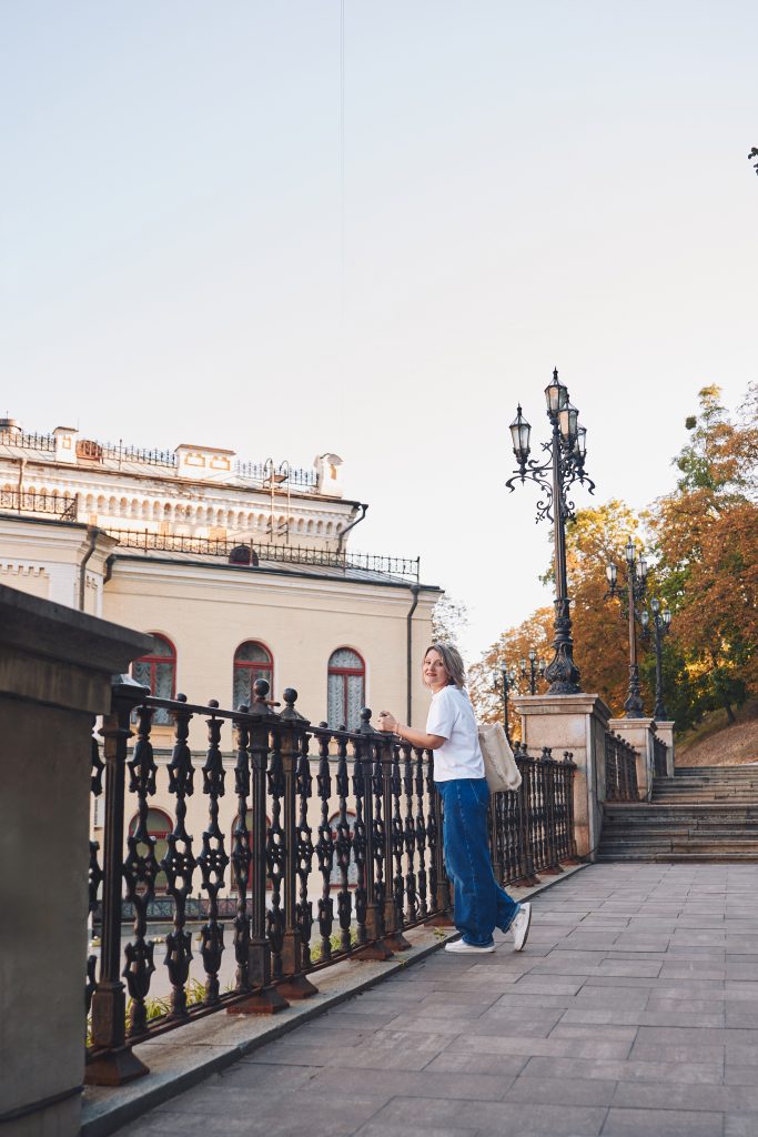 Марина стоїть біля чавунної огорожі та декоративних ліхтарів поруч зі світлою історичною будівлею.