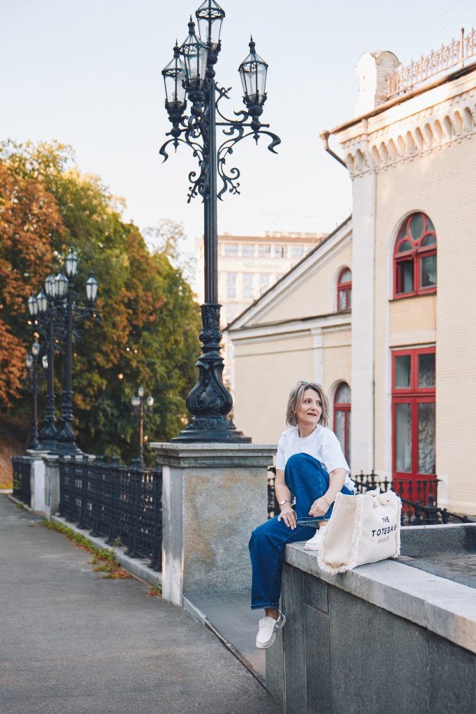 Марина сидить на кам'яному парапеті біля високого декоративного ліхтаря та світлої будівлі.