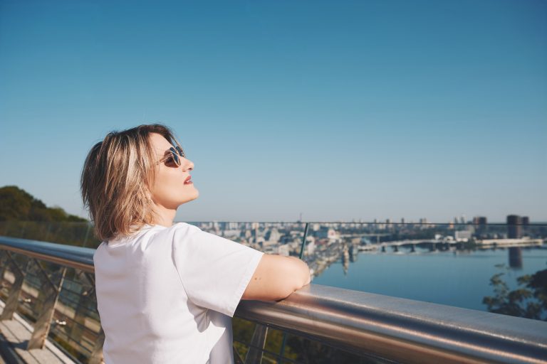 Марина в сонцезахисних окулярах спирається на огорожу Скляного мосту та дивиться на панораму Києва і Дніпро.