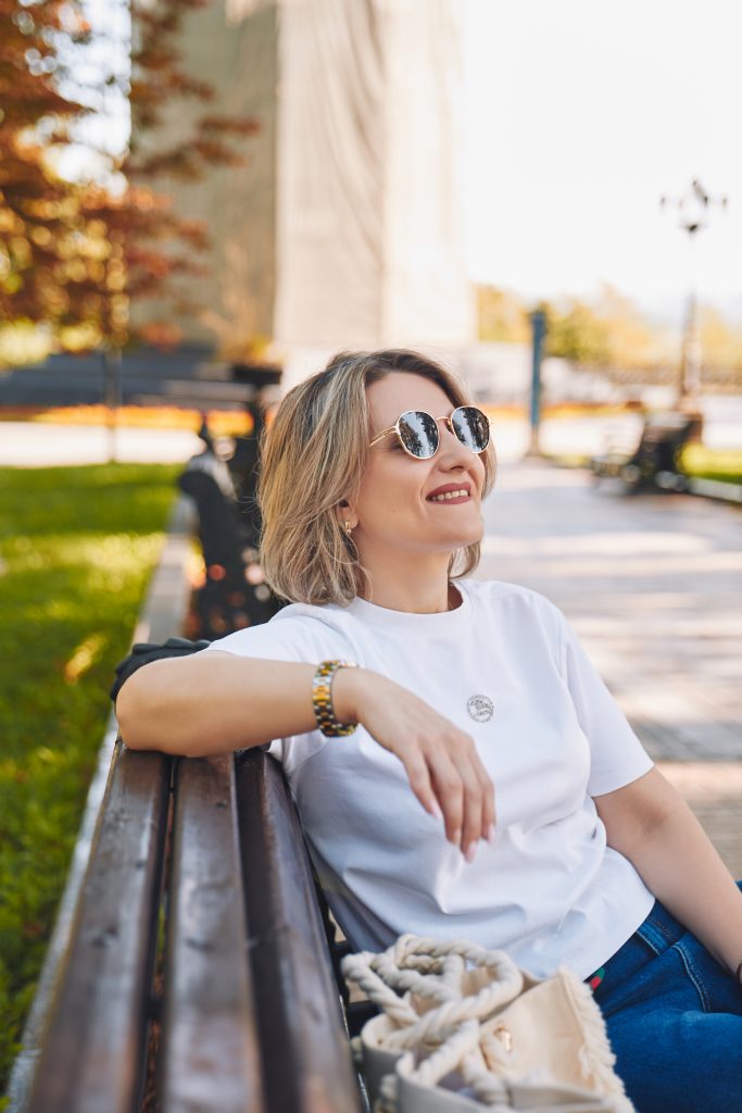 Марина в сонцезахисних окулярах сидить на парковій лавці, поруч лежить світла текстильна сумка.