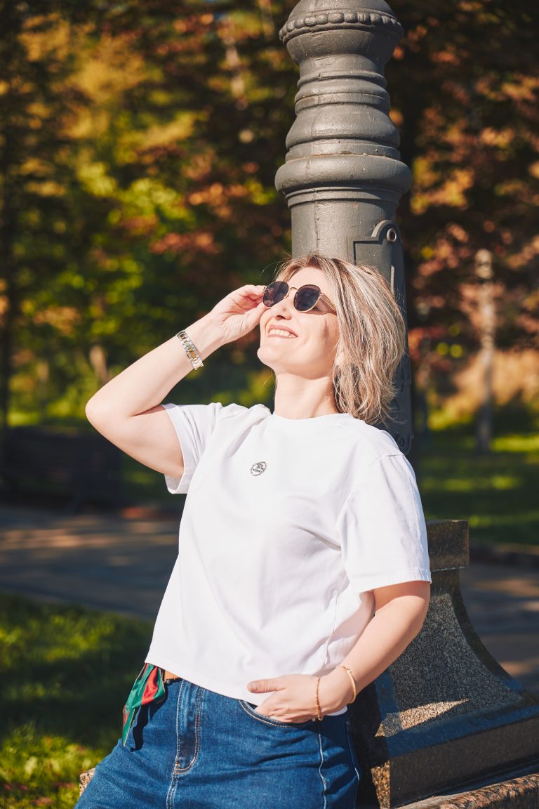 Марина в сонцезахисних окулярах усміхається біля декоративного ліхтаря на тлі зелених дерев.