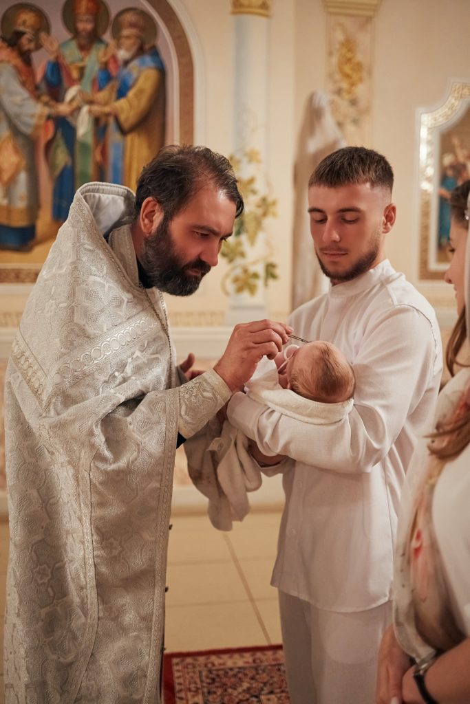 Священник торкається обличчя немовляти під час обряду, дитину тримає чоловік у білому