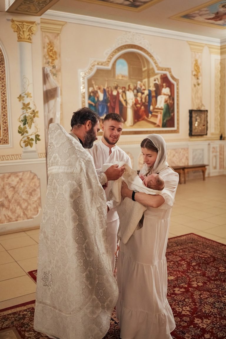 Священник передає загорнуту в рушник Емілію жінці, поруч стоїть чоловік у білому