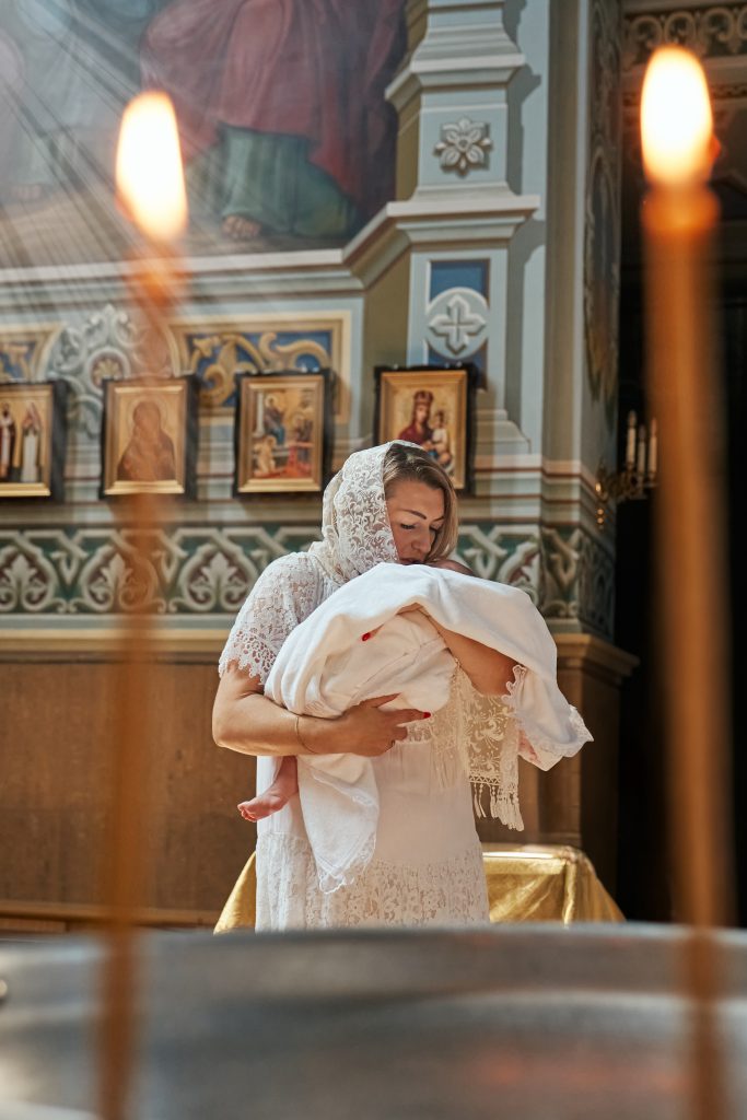 Жінка у білій мереживній хустці тримає немовля в храмі, кадр знятий крізь полум'я свічок
