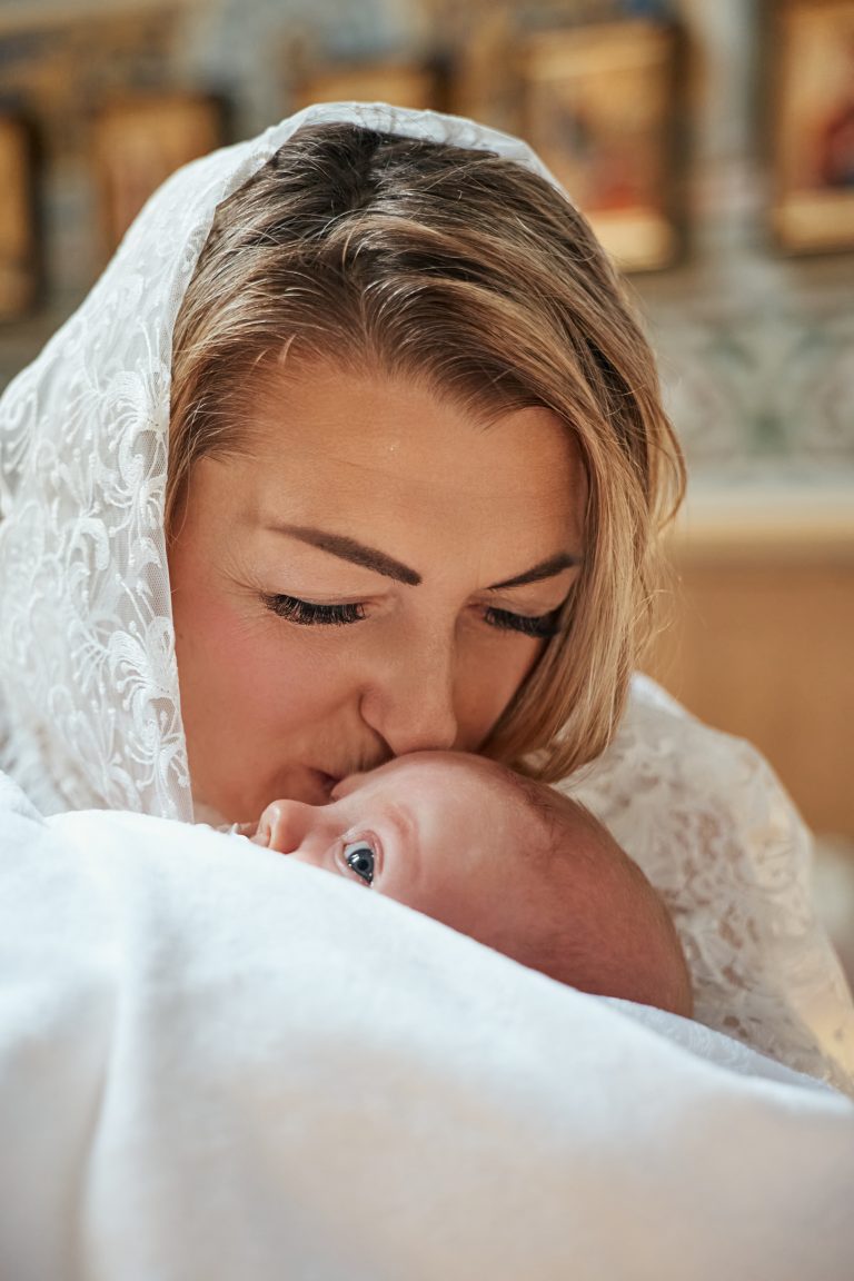 Жінка у білій мереживній хустці цілує немовля в голову під час хрещення у храмі
