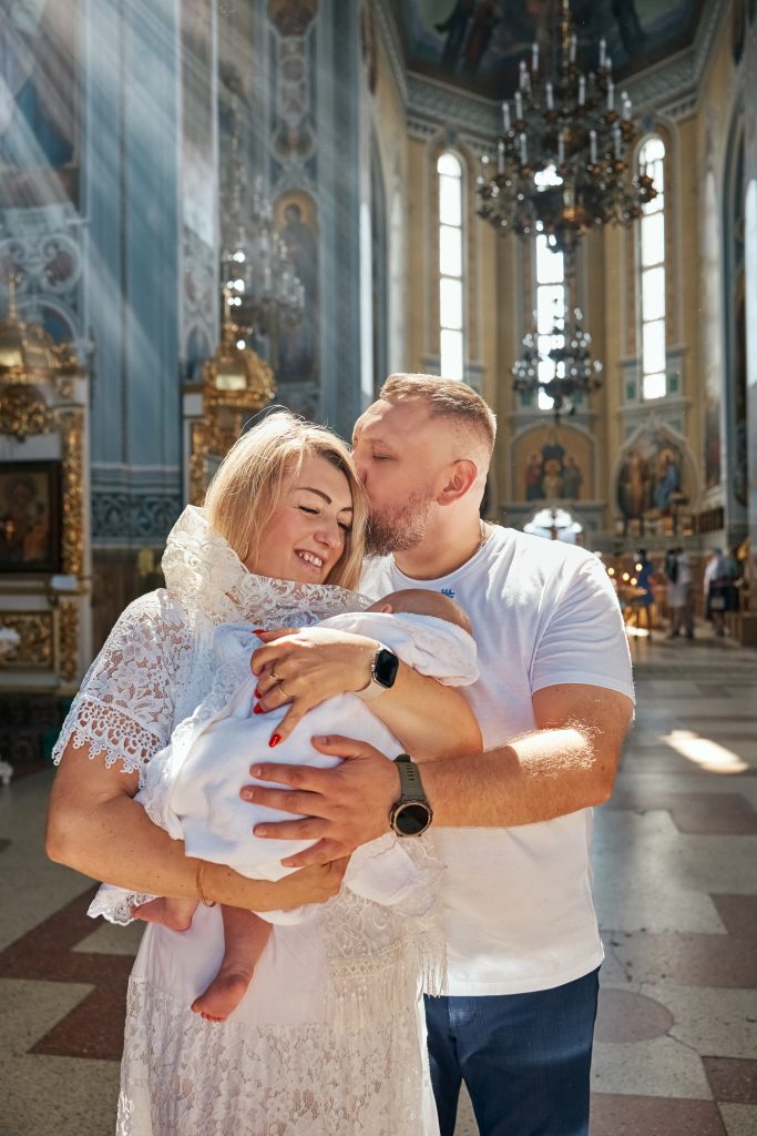 Чоловік цілує жінку в голову, поки вони разом тримають загорнуте немовля у сонячному храмі