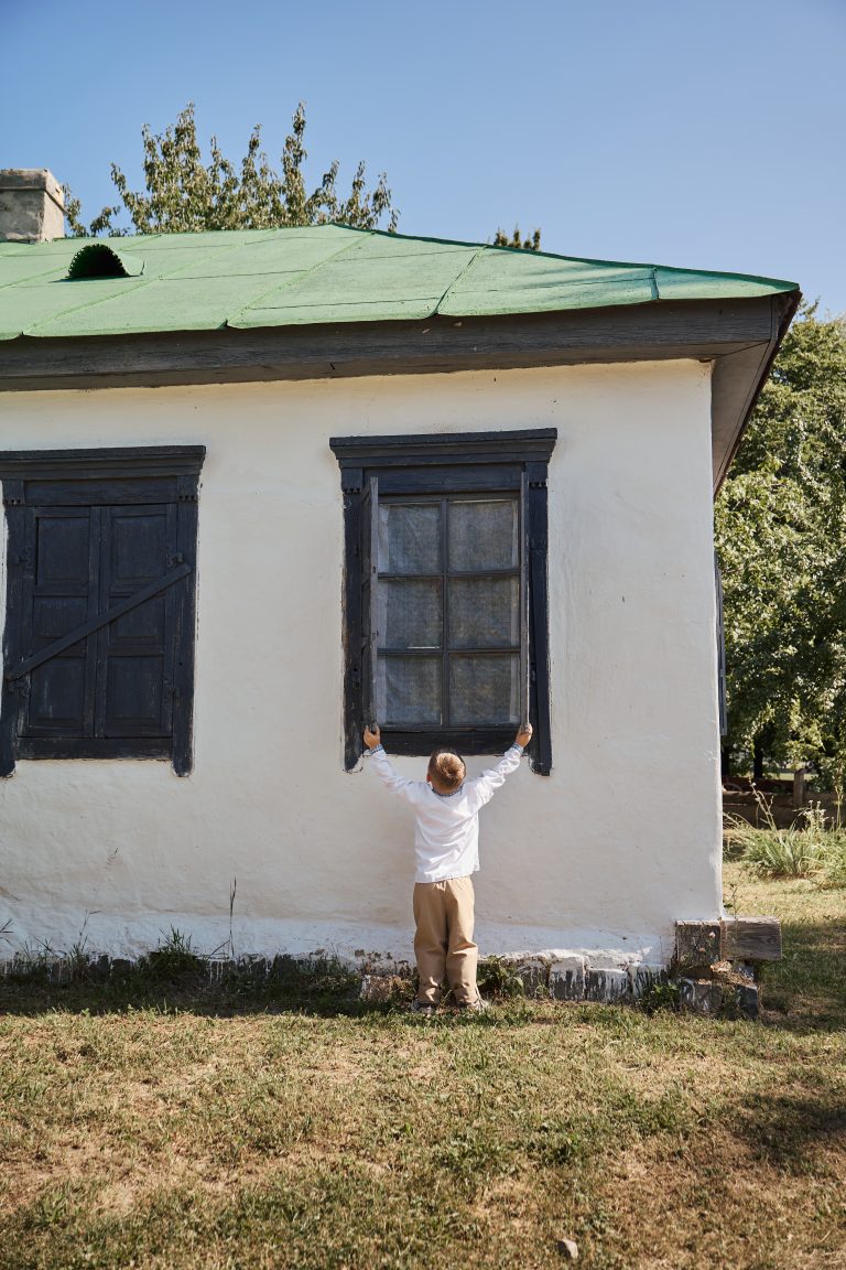 Хлопчик у вишиванці стоїть спиною біля вікна білої сільської хати