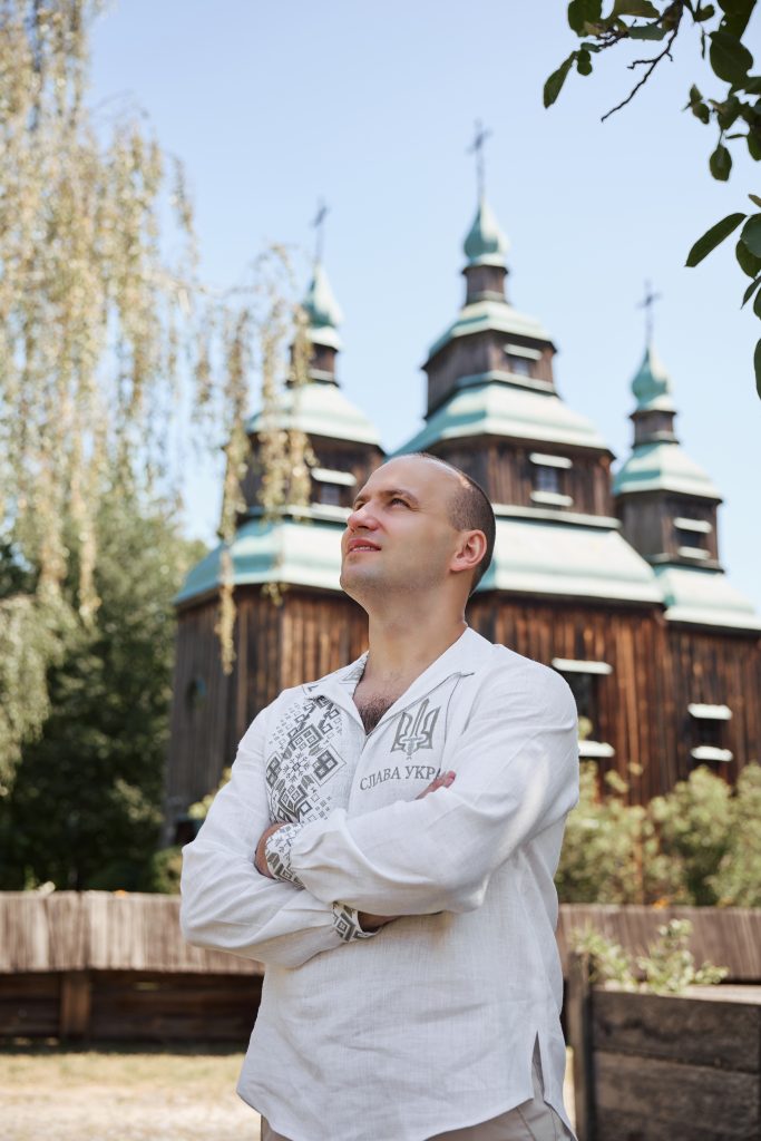 Чоловік у білій вишиванці стоїть зі схрещеними руками на тлі дерев'яного храму